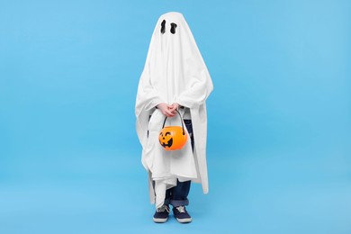 Child in white ghost costume holding pumpkin bucket on light blue background. Halloween celebration