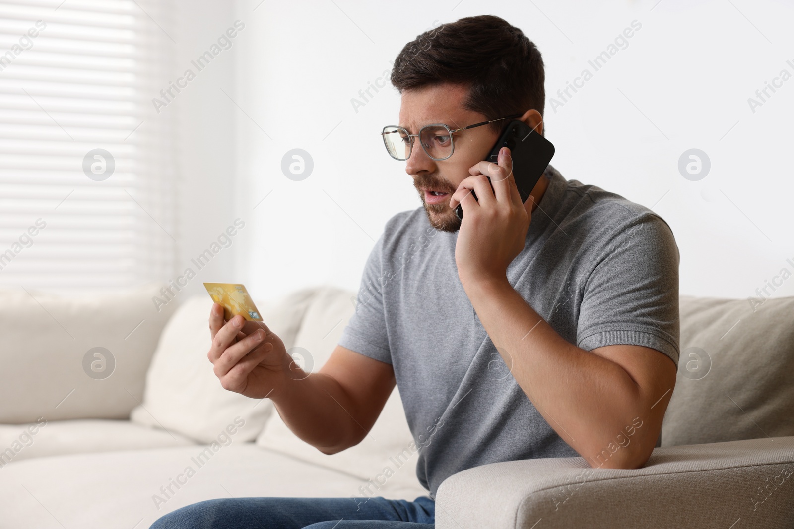 Photo of Upset man with credit card talking on smartphone at home. Be careful - fraud