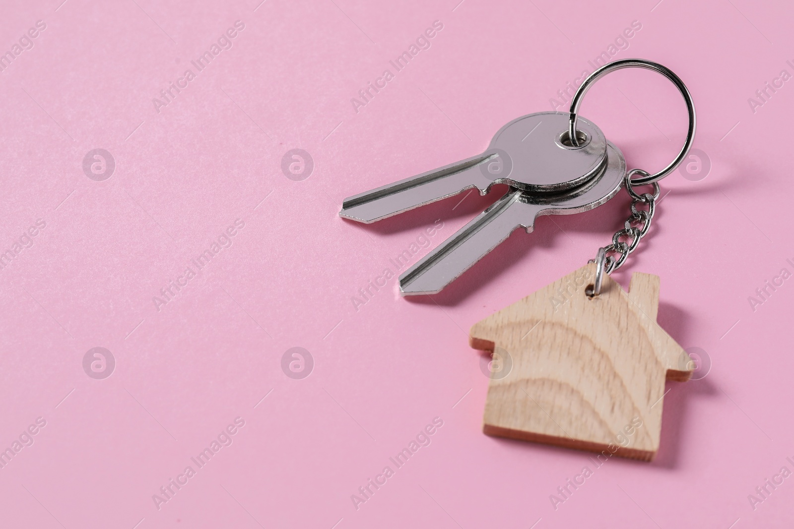 Photo of Metallic keys with wooden keychain in shape of house on pink background, space for text