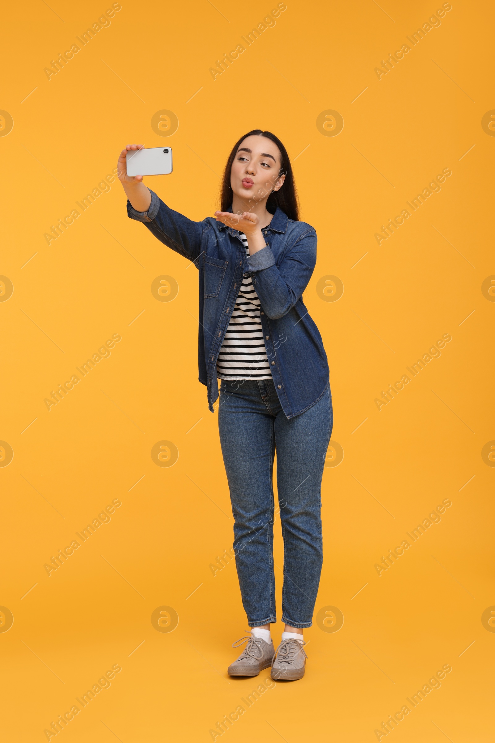 Photo of Young woman taking selfie with smartphone and blowing kiss on yellow background