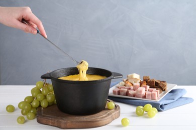 Woman dipping piece of bread into fondue pot with tasty melted cheese at white wooden table against gray background, closeup