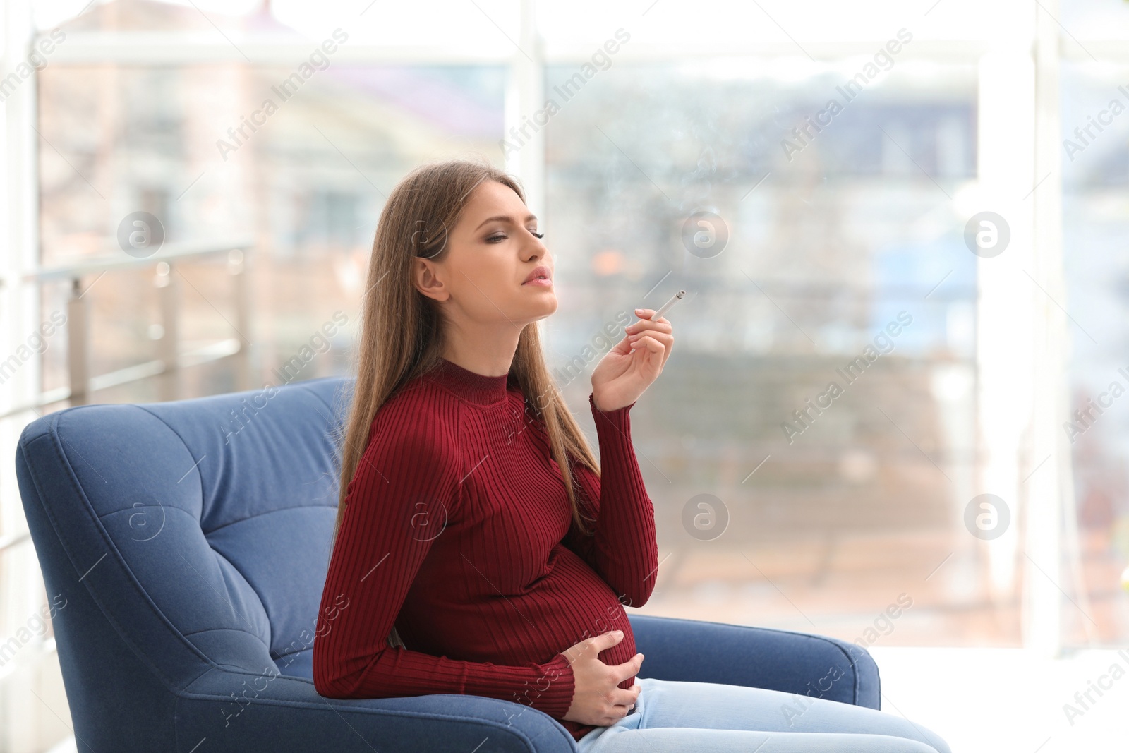 Photo of Young pregnant woman smoking cigarette at home. Harm to unborn baby