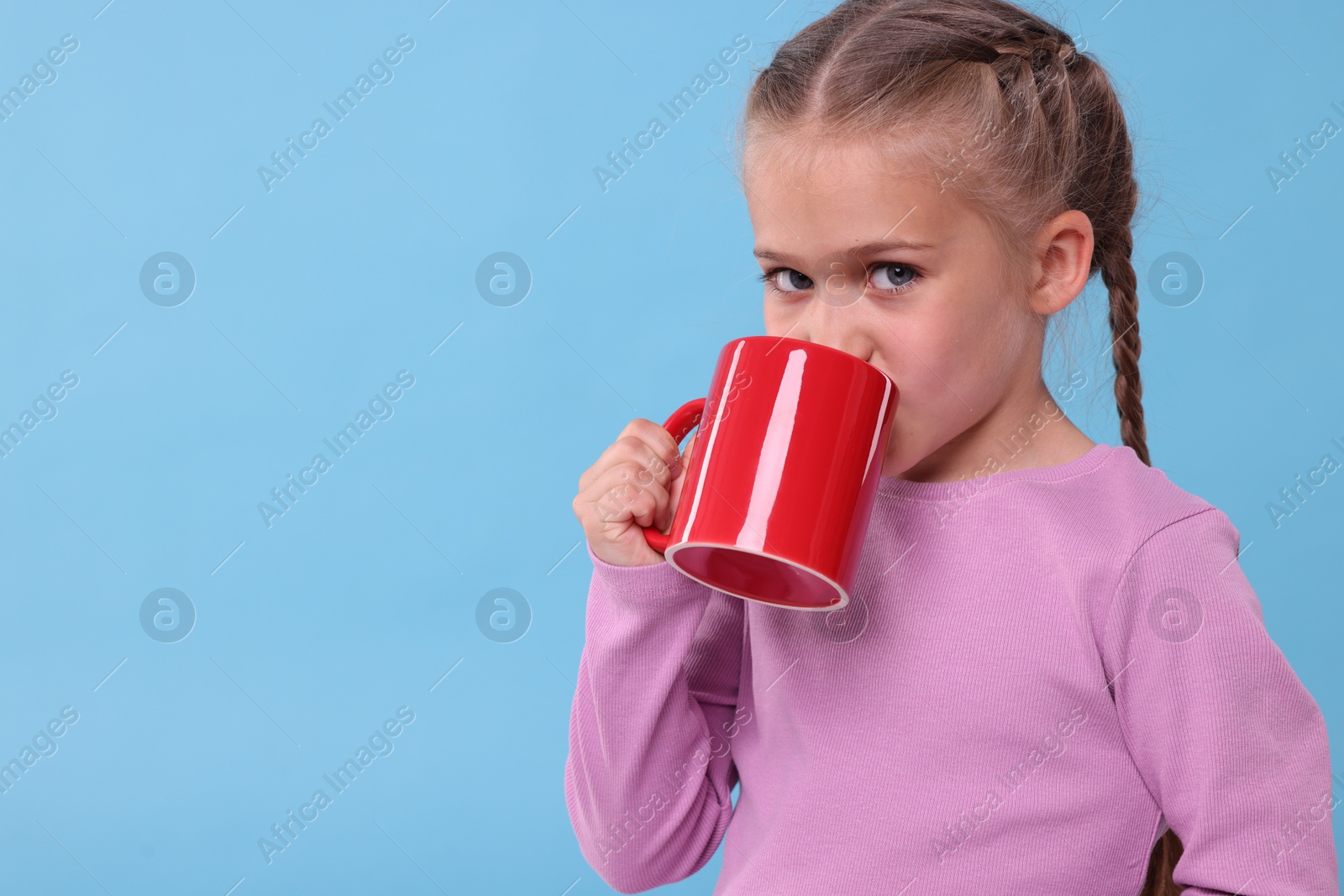 Photo of Cute girl drinking from red ceramic mug on light blue background, space for text