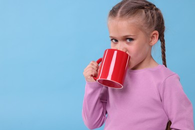 Cute girl drinking from red ceramic mug on light blue background, space for text