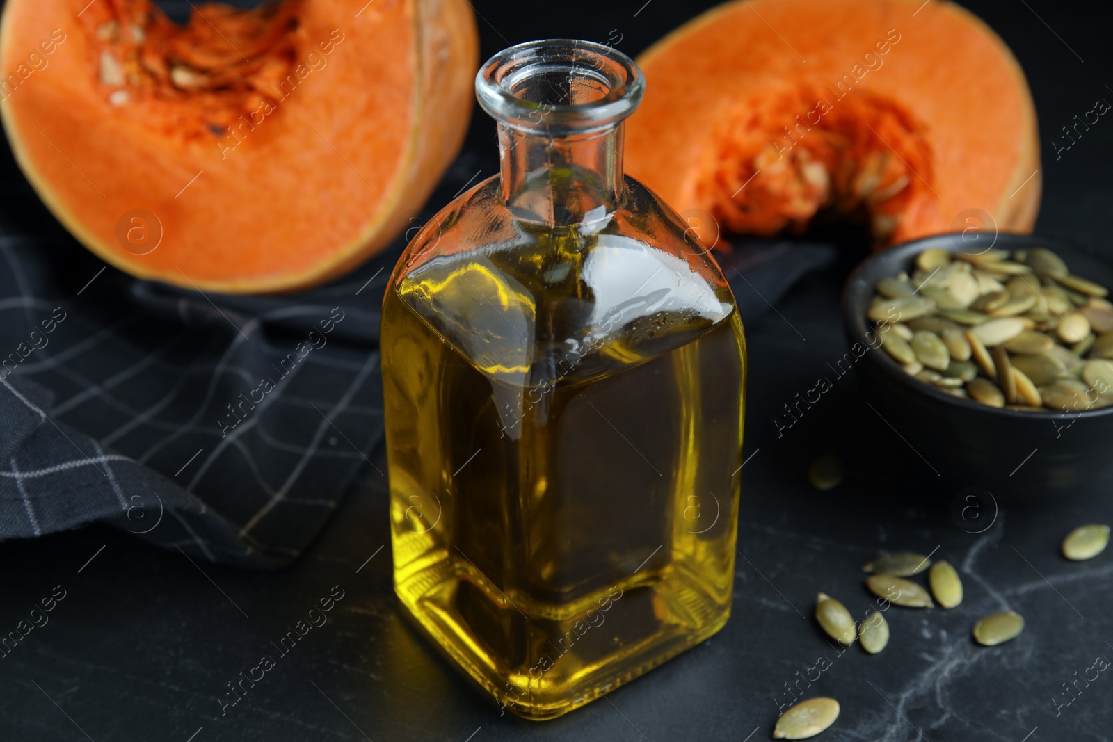 Photo of Glass bottle of oil with cut pumpkin and seeds on black table, closeup