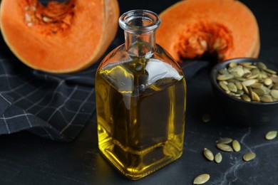Photo of Glass bottle of oil with cut pumpkin and seeds on black table, closeup