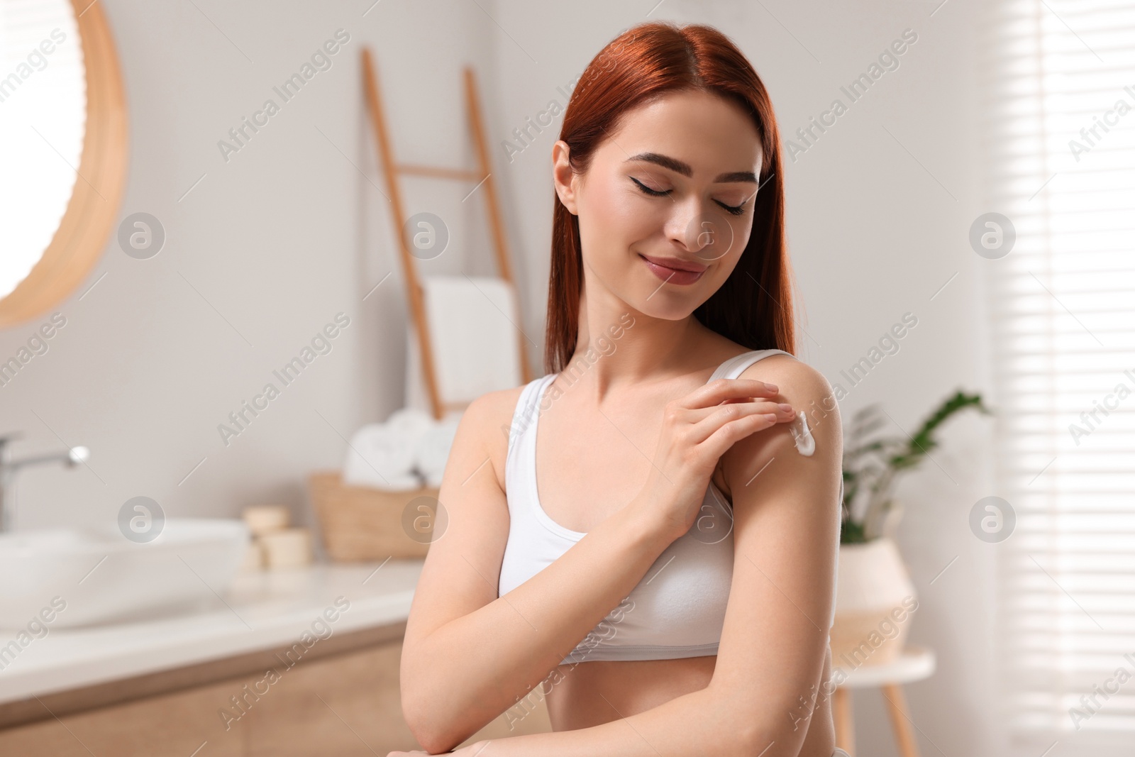 Photo of Beautiful young woman applying body cream onto shoulder in bathroom, space for text