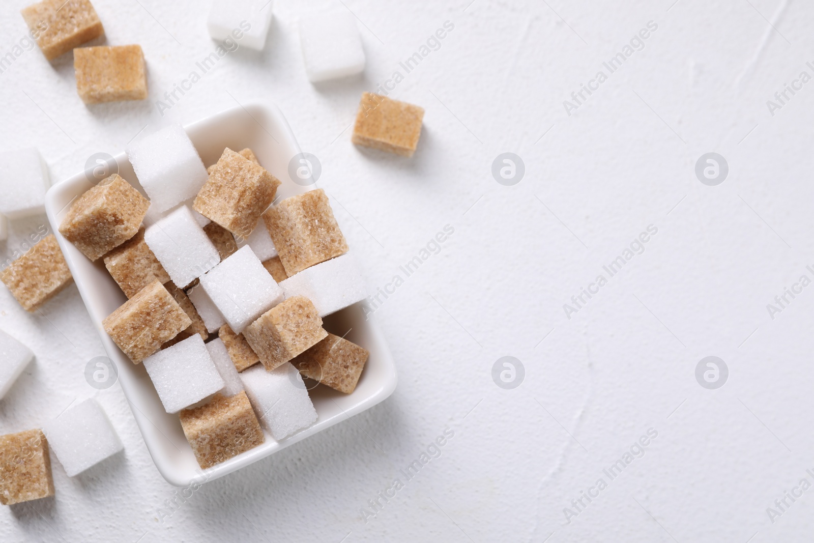 Photo of Different sugar cubes in bowl on white table, top view. Space for text