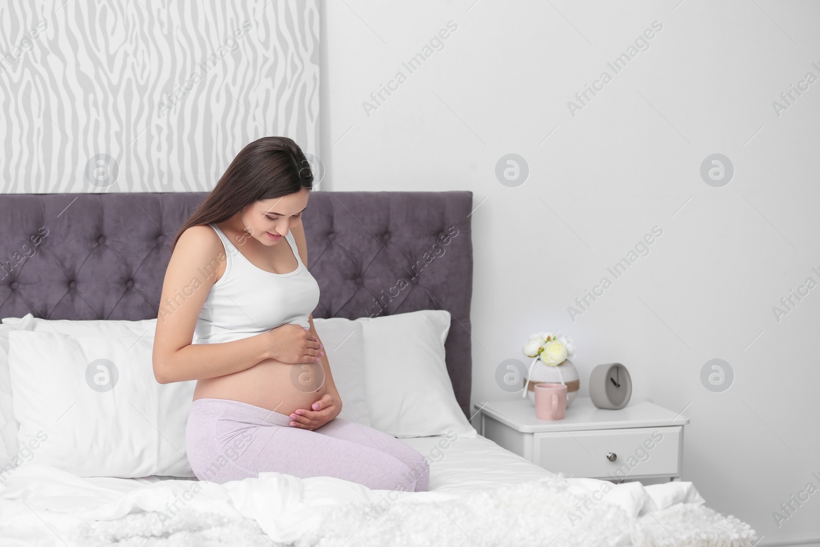 Photo of Young beautiful pregnant woman sitting on bed and touching her belly at home