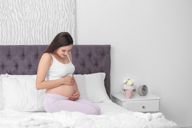 Young beautiful pregnant woman sitting on bed and touching her belly at home