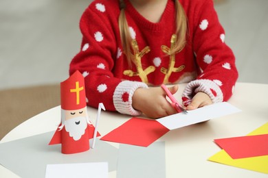 Photo of Cute little girl cutting paper at table with Saint Nicholas toy indoors, closeup