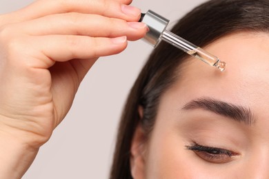 Photo of Young woman applying essential oil onto face on light grey background, closeup