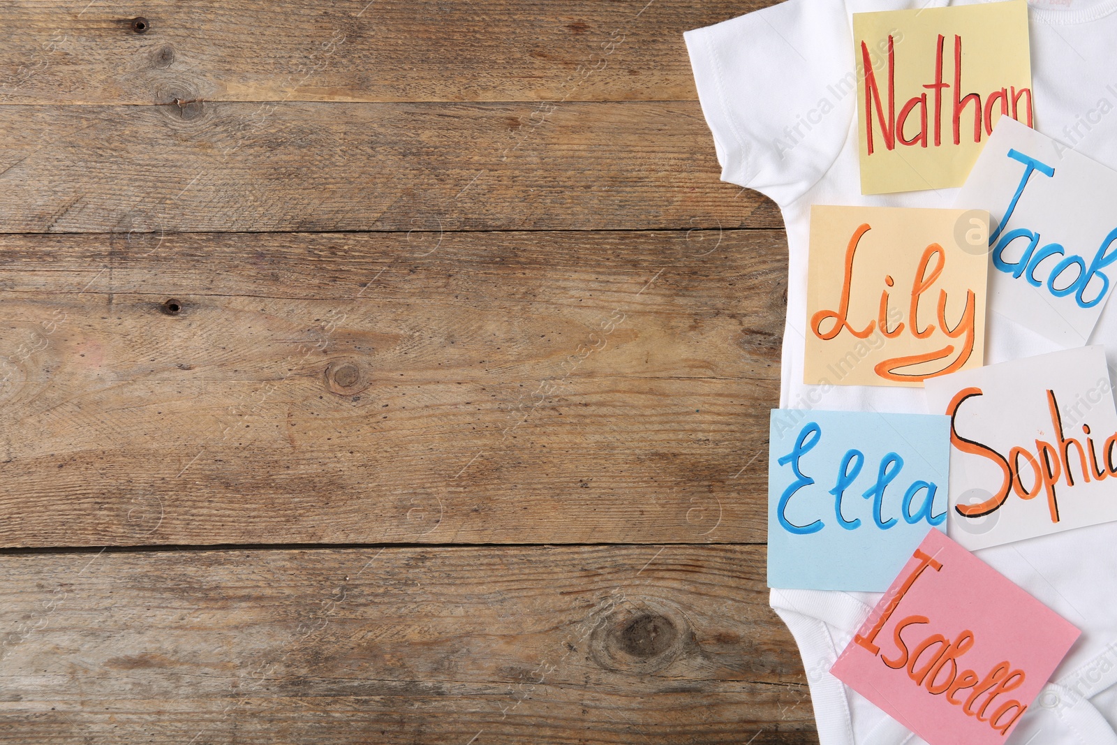 Photo of Paper notes with different baby names and child's bodysuit on wooden table, top view. Space for text