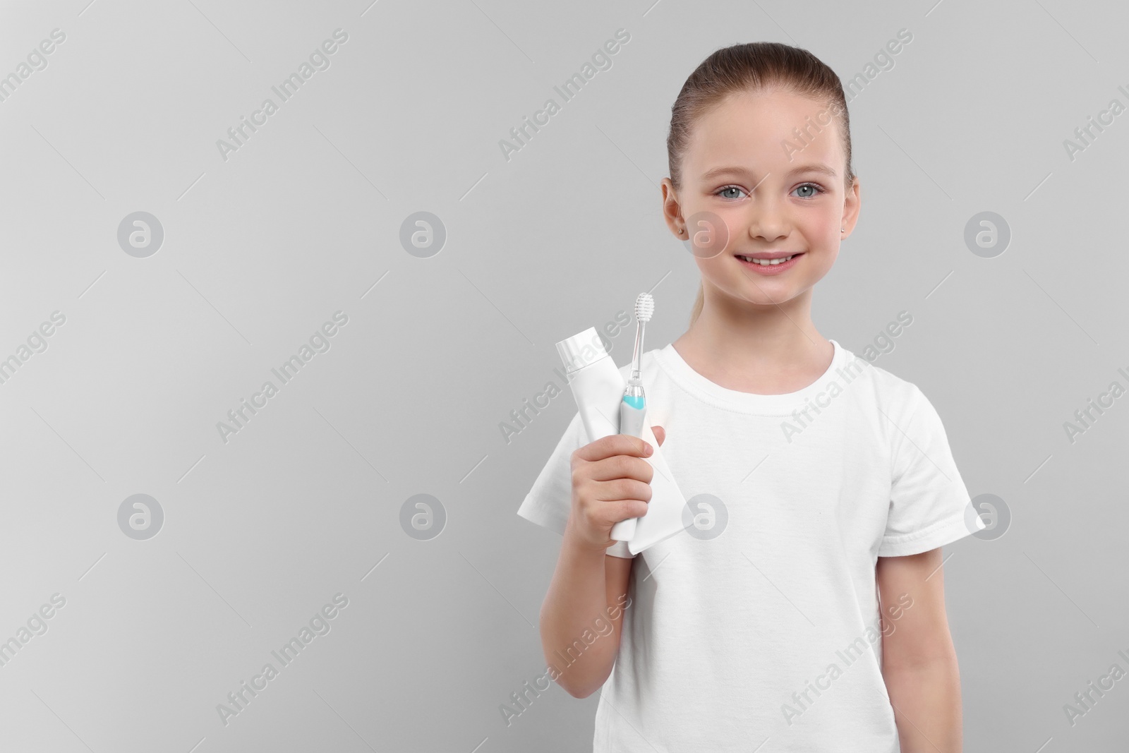 Photo of Happy girl holding electric toothbrush and tube of toothpaste on light grey background. Space for text