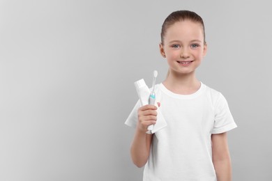 Happy girl holding electric toothbrush and tube of toothpaste on light grey background. Space for text