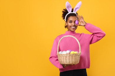 Photo of Happy African American woman in bunny ears headband covering eye with Easter egg on orange background, space for text