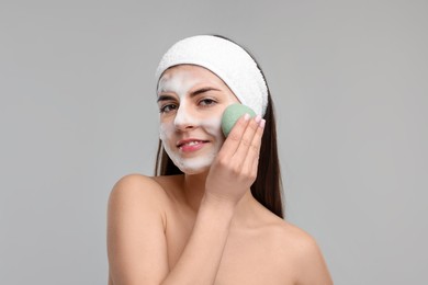 Photo of Young woman with headband washing her face using sponge on light grey background