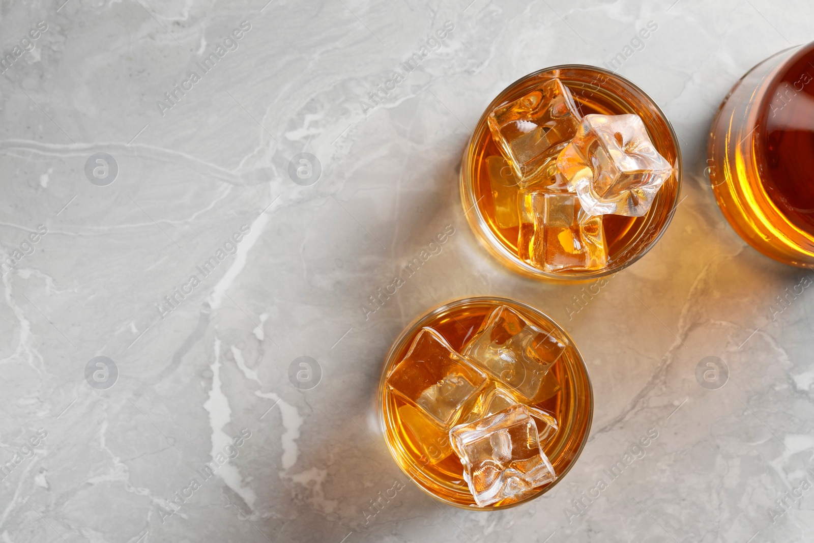 Photo of Whiskey with ice cubes in glasses and bottle on light grey textured table, flat lay. Space for text