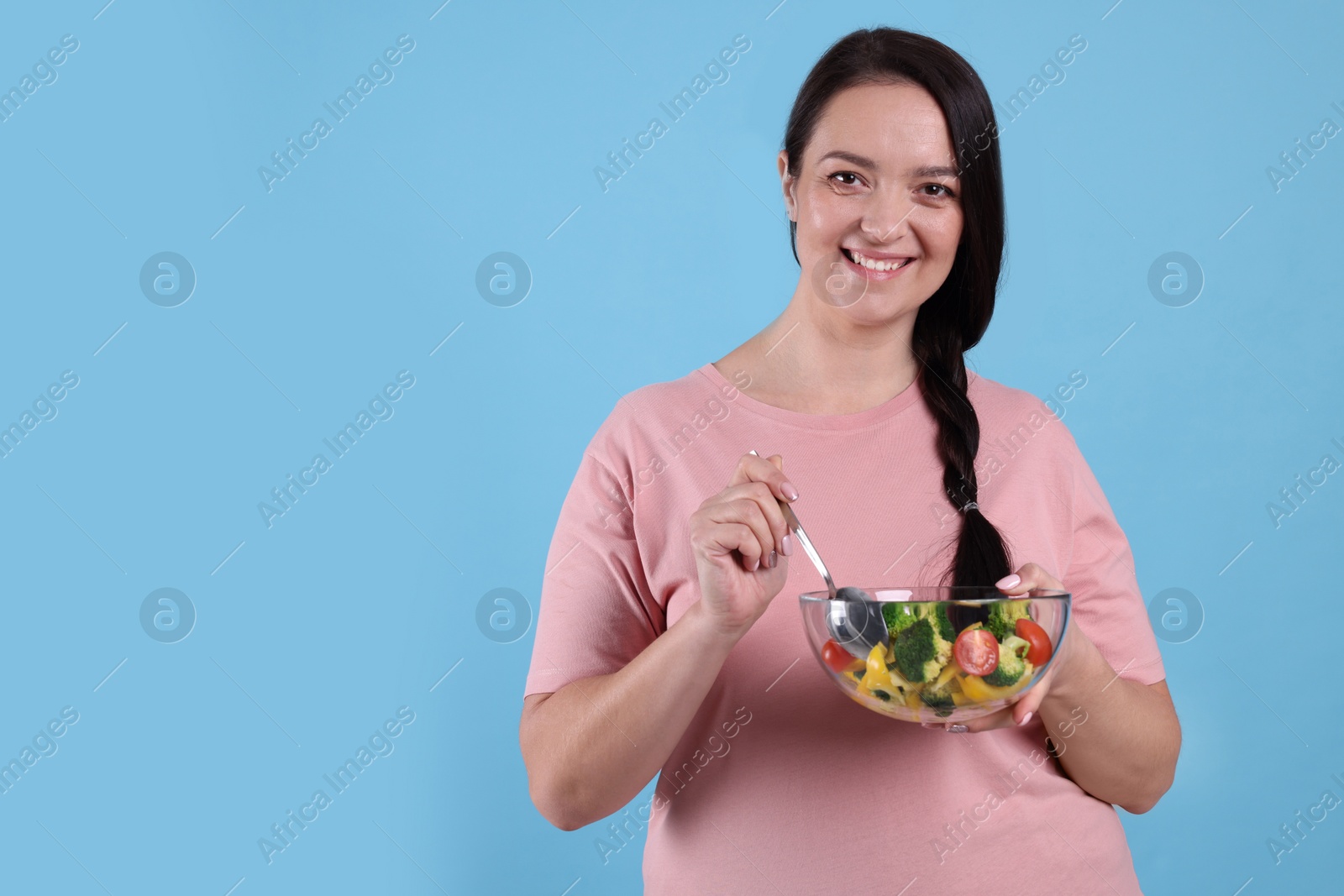 Photo of Beautiful overweight woman eating salad on light blue background, space for text. Healthy diet