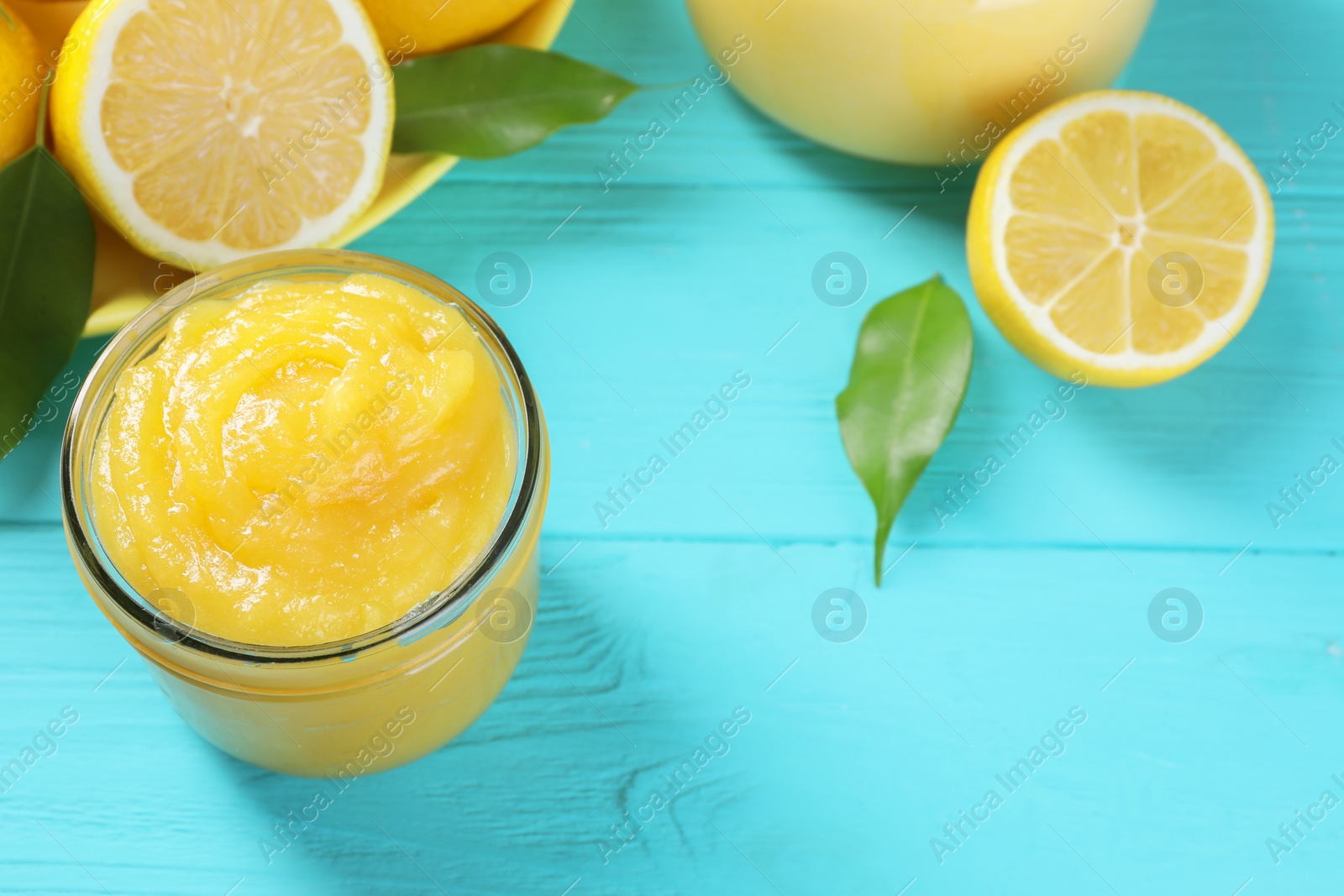 Photo of Delicious lemon curd in glass jar, fresh citrus fruit and green leaves on light blue wooden table