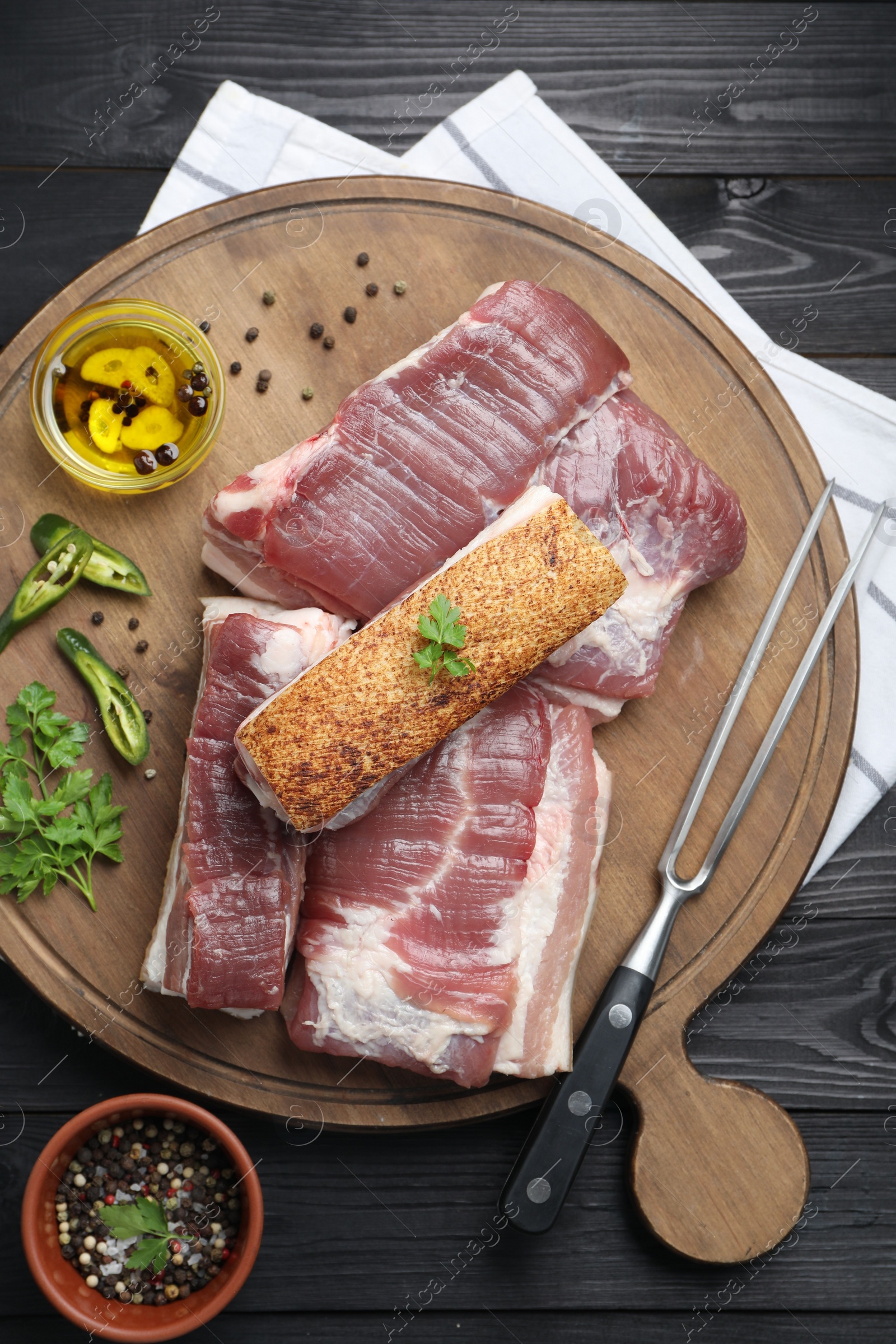 Photo of Pieces of raw pork belly, chili pepper, peppercorns, oil and parsley on black wooden table, top view