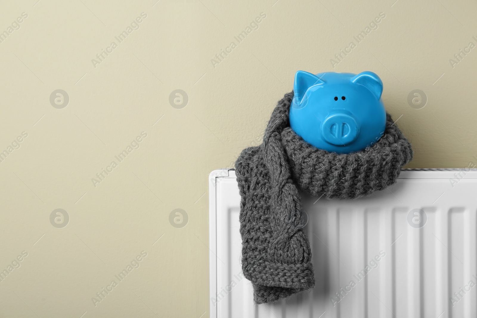 Photo of Piggy bank wrapped in scarf on heating radiator against beige background, space for text