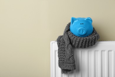 Photo of Piggy bank wrapped in scarf on heating radiator against beige background, space for text