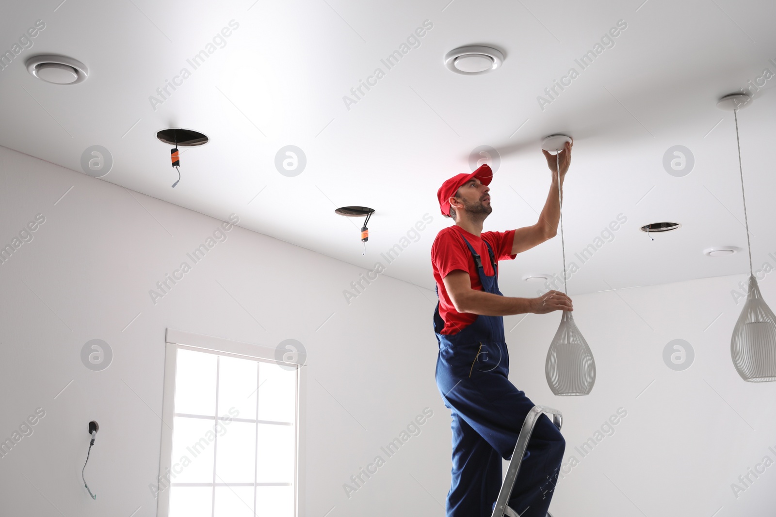 Photo of Worker installing lamp on stretch ceiling indoors