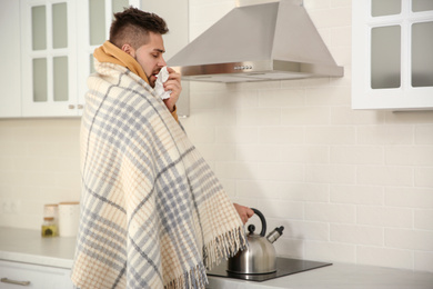 Sick young man putting heating kettle in kitchen. Influenza virus