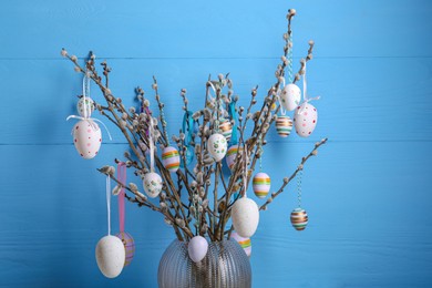 Photo of Beautiful willow branches with painted eggs in vase on light blue wooden background. Easter decor