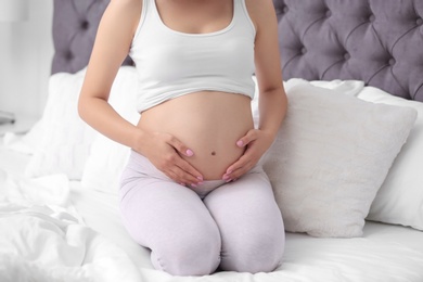 Photo of Young pregnant woman sitting on bed and touching her belly at home, closeup