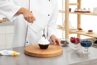 Photo of Female pastry chef pouring chocolate sauce onto cupcake at table in kitchen, closeup