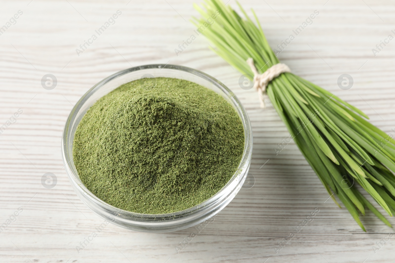 Photo of Wheat grass powder in glass bowl and fresh sprouts on white wooden table, closeup