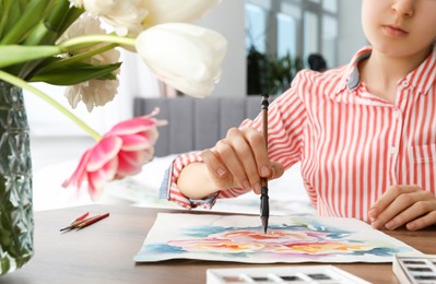 Photo of Woman painting flowers with watercolor at wooden table indoors, closeup. Creative artwork