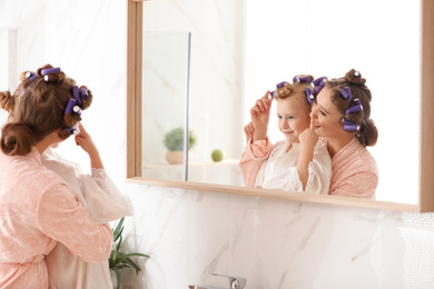 Photo of Happy mother and daughter with curlers near mirror in bathroom