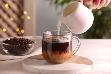 Woman pouring milk into cup with aromatic coffee at white wooden table, closeup