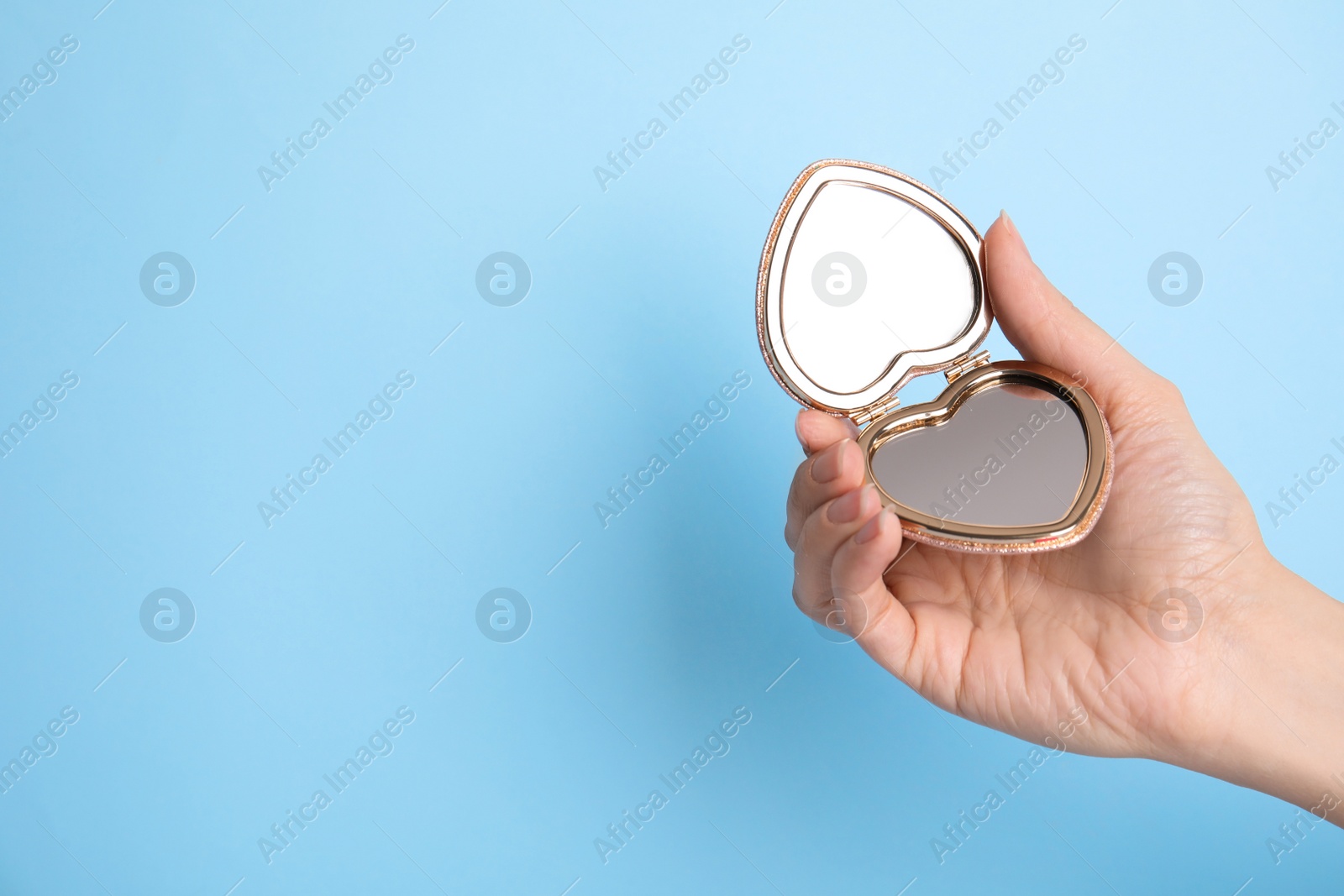 Photo of Woman holding stylish heart shaped cosmetic pocket mirror on light blue background, closeup. Space for text