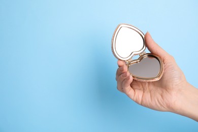 Woman holding stylish heart shaped cosmetic pocket mirror on light blue background, closeup. Space for text