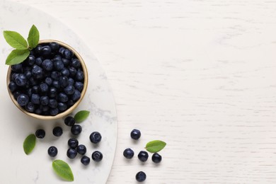 Photo of Bowl of tasty fresh bilberries with green leaves on white wooden table, flat lay. Space for text