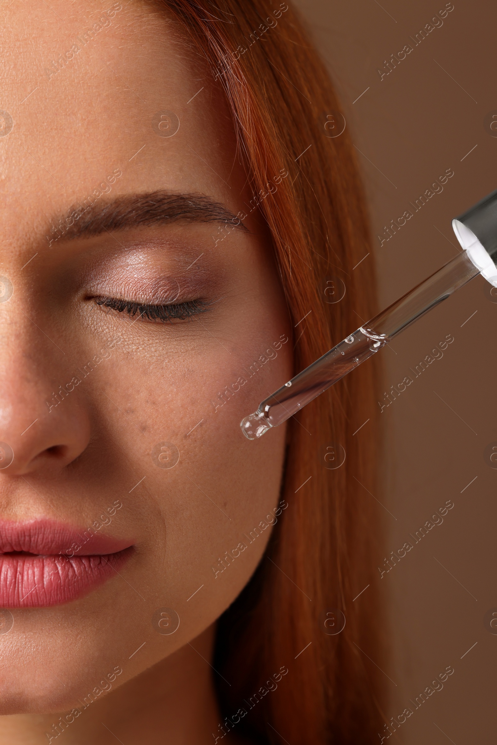 Photo of Beautiful young woman applying cosmetic serum onto her face on brown background, closeup