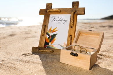 Photo of Beautiful wedding invitation and wooden box with gold rings on sandy beach