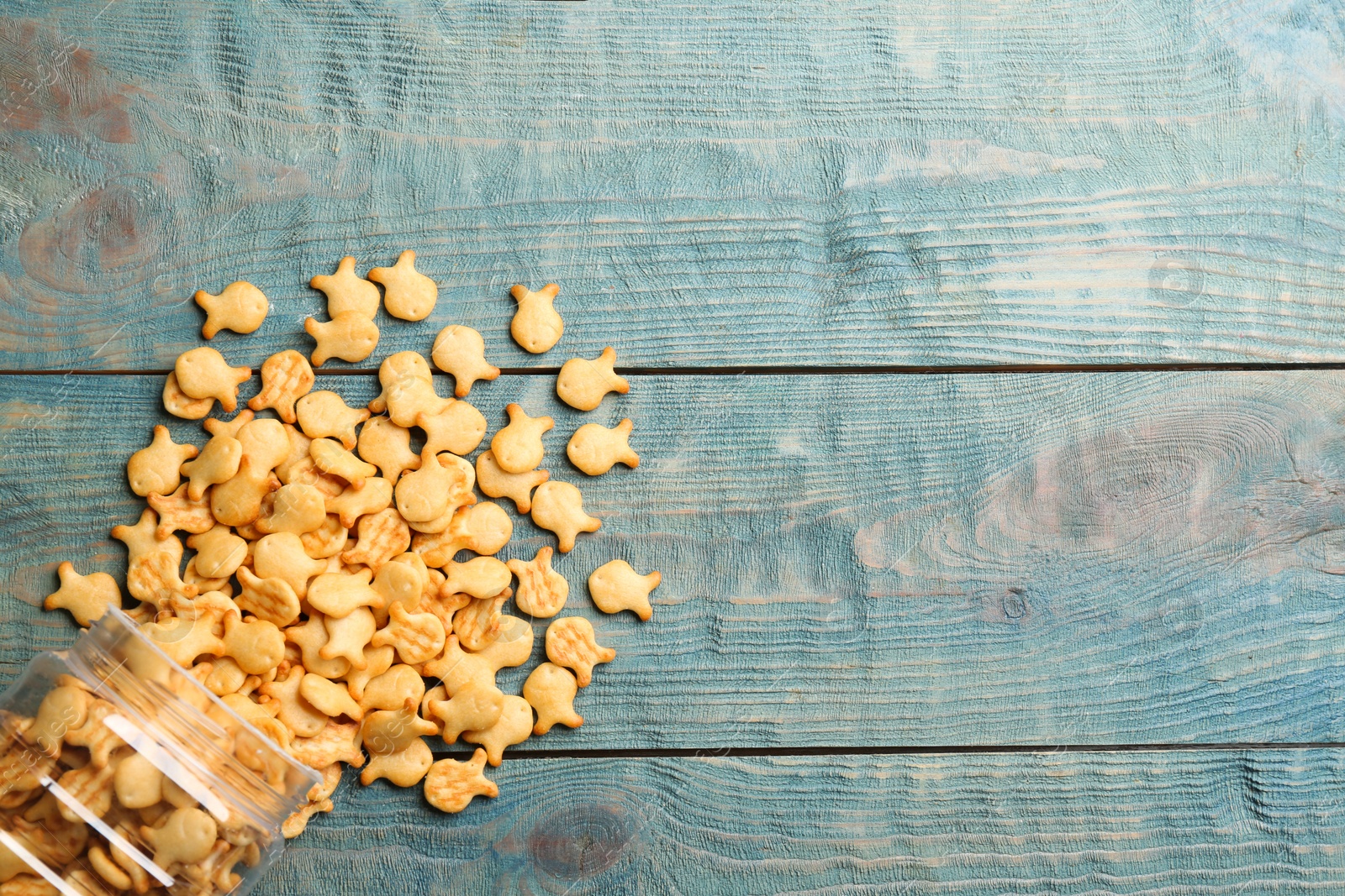 Photo of Overturned jar with goldfish crackers on blue wooden table, flat lay. Space for text
