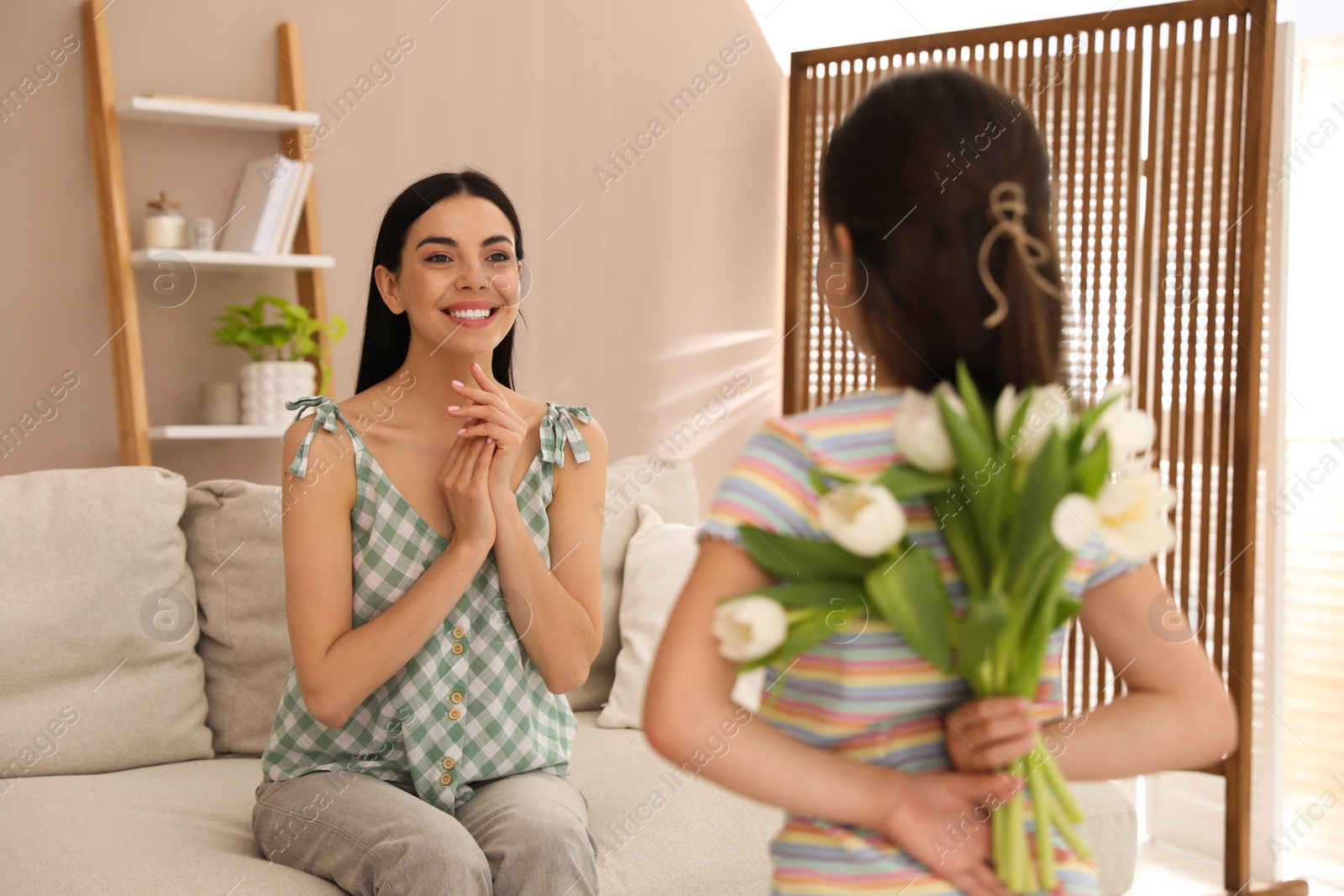 Photo of Little girl hiding tulip bouquet for mom at home. Happy Mother's Day