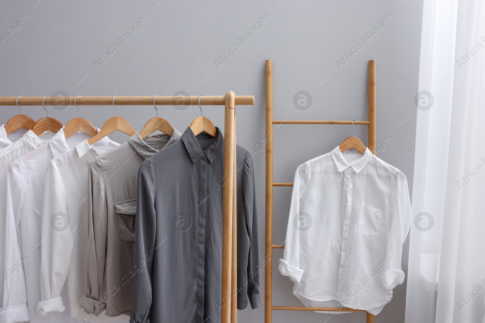 Photo of Different stylish shirts hanging on ladder and rack near grey wall in room. Organizing clothes