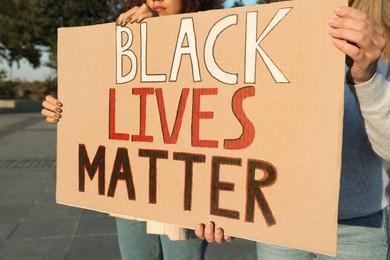 Photo of Women holding sign with phrase Black Lives Matter outdoors, closeup. Racism concept