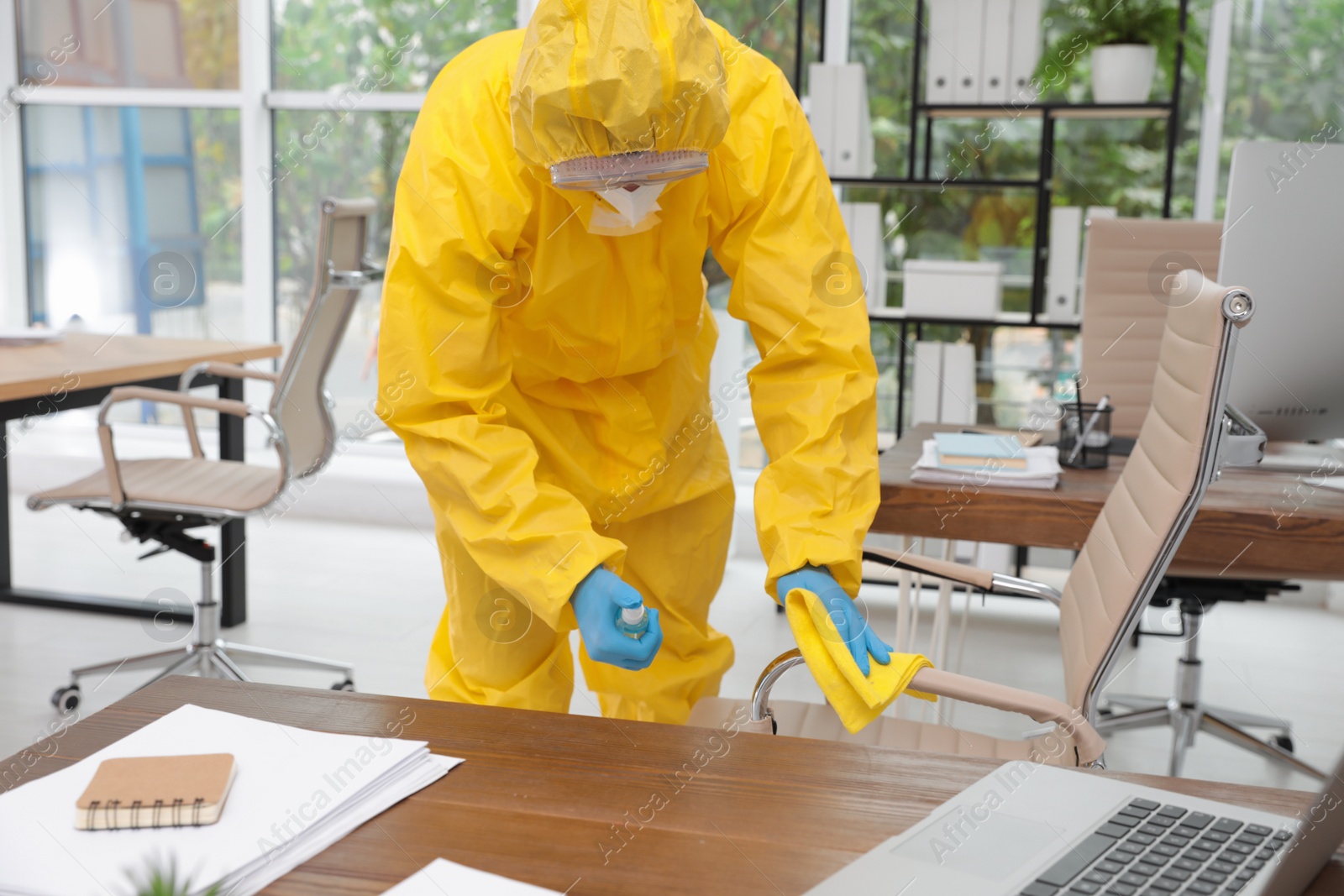 Photo of Janitor in protective suit disinfecting office furniture to prevent spreading of COVID-19