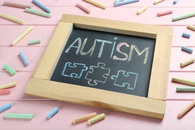 Blackboard with word Autism and drawn jigsaw puzzle pieces surrounded by colorful chalks on pink wooden background