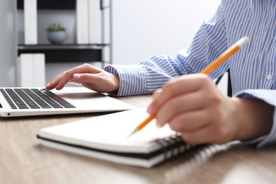 Woman with notebook working on laptop at wooden table in office, closeup