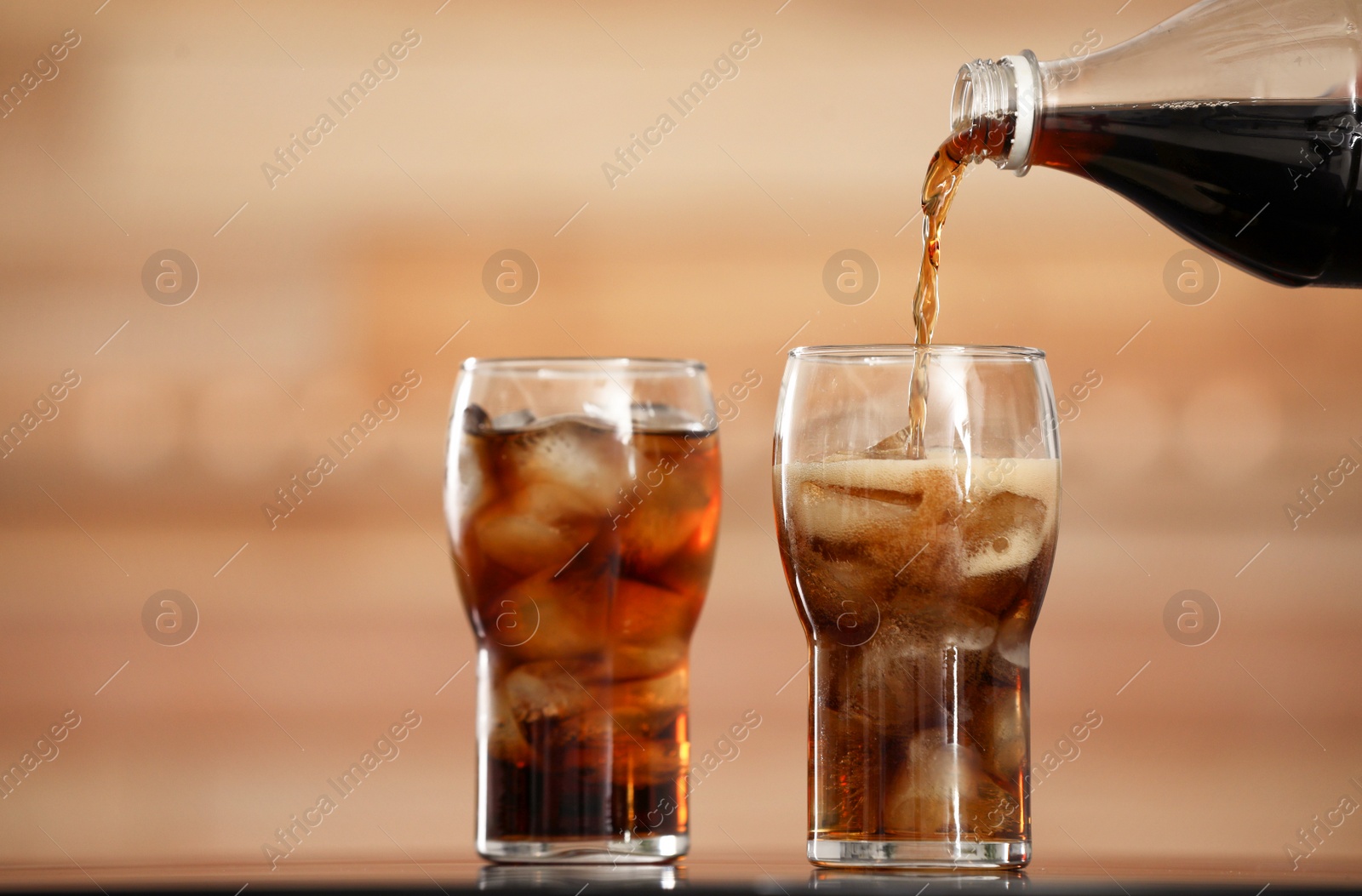 Photo of Pouring cola from bottle into glass with ice cubes on table against blurred background
