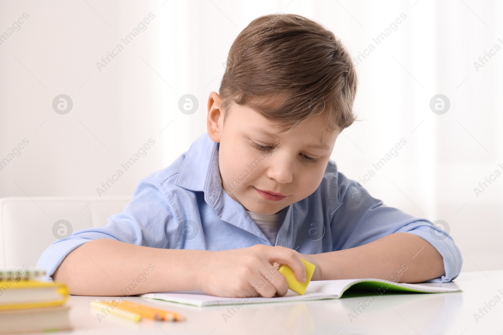 Photo of Little boy erasing mistake in his notebook at white desk indoors
